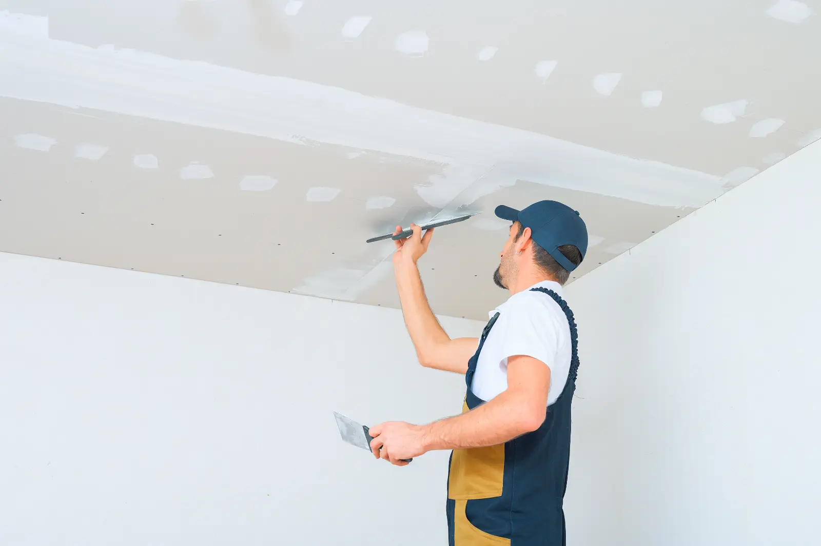 Drywall Installation. A uniformed worker applies putty to the drywall ceiling. Putty of joints of drywall sheets